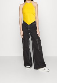 Woman wearing a yellow halter top, black wide-leg cargo pants with white stitching, and silver platform sneakers, standing against a plain background.