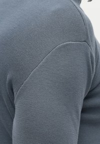 Tuvplānā bilde ar cilvēka plecu, kurš valkā teksturētu, ribotu, zilu pelēku adītu džemperi.