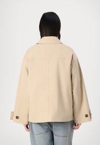 Beige, oversized jakke lavet af et glat materiale. Har en bred krave og store knapper ved manchetten. Bæres over blå denim.