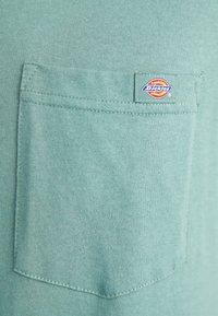 Mátově zelené bavlněné tričko s přední kapsou. Obsahuje obdélníkovou nášivku loga Dickies přišitou nad kapsou. Hladká textura.