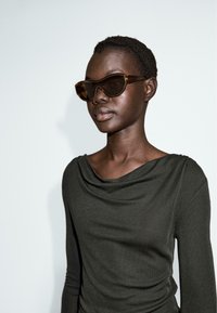 Occhiali da sole tartarugati con design angolare indossati da un modello in un top a maniche lunghe drappeggiato di colore verde scuro su uno sfondo chiaro.