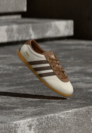 Λευκό και καφέ αθλητικό παπούτσι Adidas Gazelle με σόλα από καουτσούκ να αιωρείται πάνω από πέτρινα σκαλοπάτια και έδαφος σε εξωτερικό χώρο.