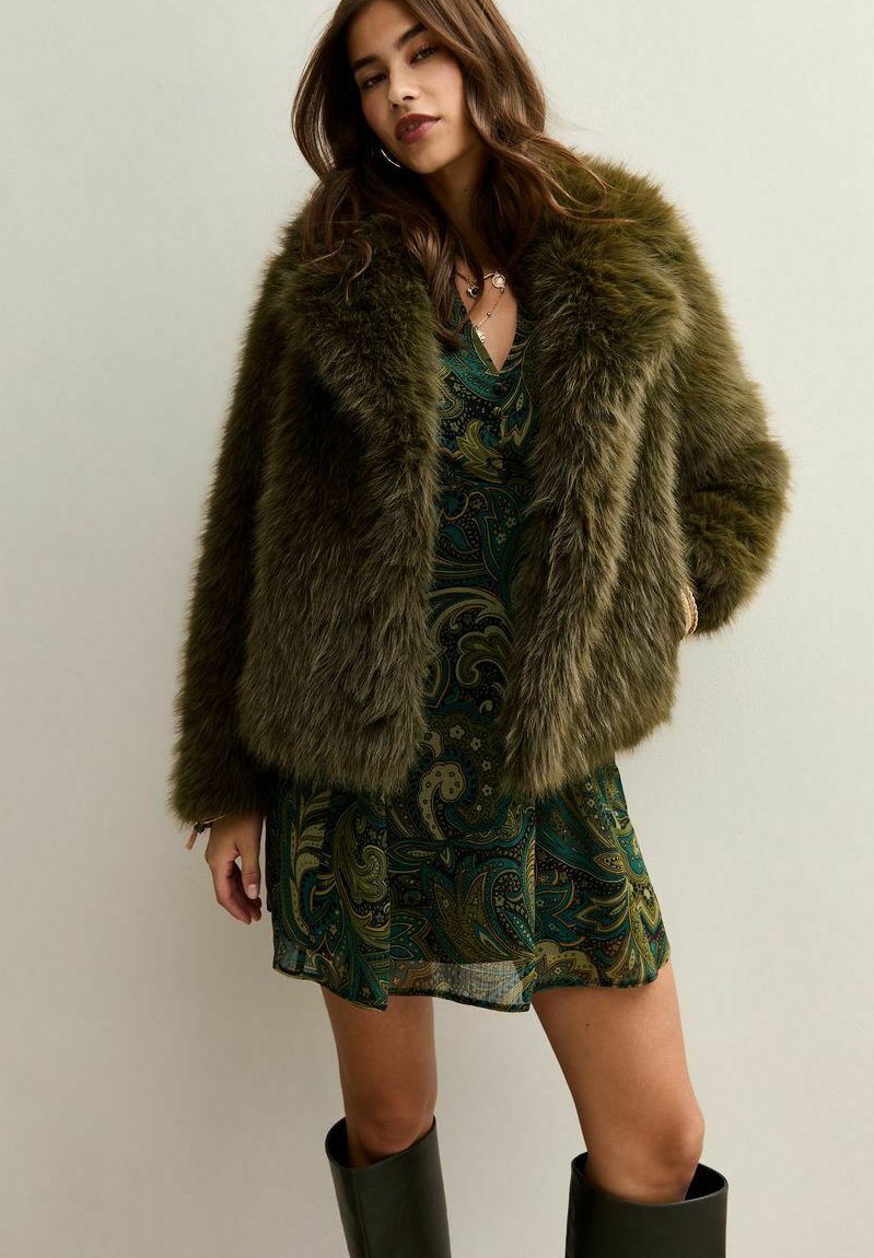 Kobieta ubrana w ciemnozieloną kurtkę z futra syntetycznego na wzorzystej zielonej sukience w paisley oraz czarnych kozakach do kolan, stojąca na jednolitym tle.
