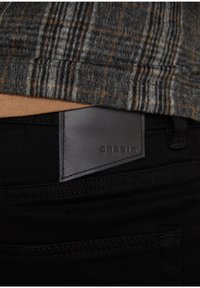 Jeans de denim negro presentan una etiqueta de cuero marrón con "CHASIM" grabado, ubicada justo por encima de la cinturilla, acompañados de una camisa de cuadros gris.