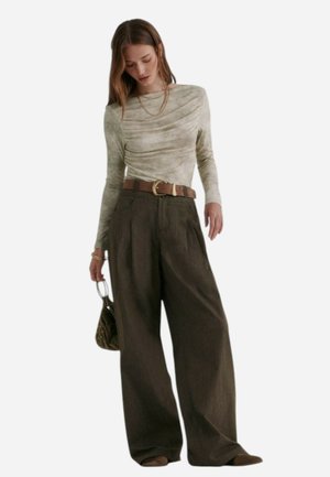 Top fitted a maniche lunghe di colore verde chiaro con scollatura drappeggiata, abbinato a pantaloni marroni a gamba larga e una cintura marrone. Tiene una borsa a mano di piccole dimensioni.