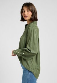 Blusa a maniche lunghe verde oliva con righe verticali, polsini con bottoni e una vestibilità morbida, caratterizzata da un colletto a scollo a V e un tessuto morbido e liscio.