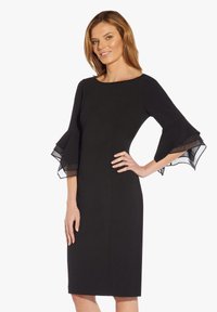 Robe noire avec une silhouette ajustée, dotée de manches transparentes évasées. Texture lisse avec une coupe au niveau du genou et un col arrondi.