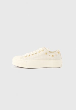 Zapatilla baja color crema con suela gruesa, ojales dorados y pequeños tachuelas doradas en forma floral y redonda en el panel lateral.