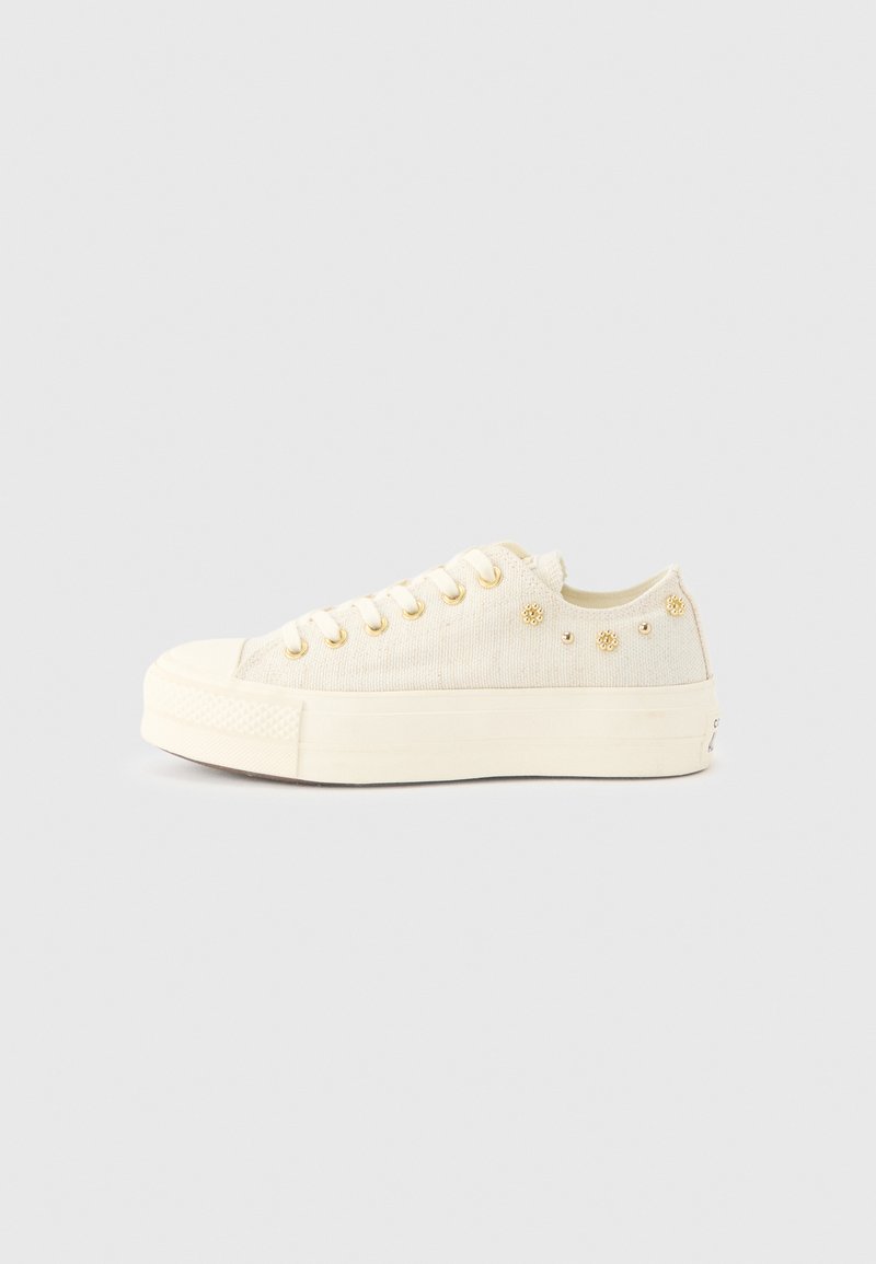 Zapatilla baja color crema con suela gruesa, ojales dorados y pequeños tachuelas doradas en forma floral y redonda en el panel lateral.