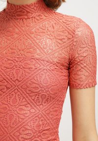 Top maniche corte in pizzo corallo, caratterizzato da intricate fantasie floreali e un colletto alto. Il tessuto testurizzato presenta un delicato intreccio aperto.