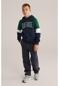 Jūras spēku kapuce ar zaļiem un baltiem akcentiem, uz kuras ir rakstīts "BLUE RIDGE"; kopā ar pelēkām kordura biksēm un pelēkām kedām.