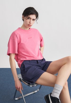 Persona con el cabello corto y oscuro, sentada en una silla de metal, vistiendo una camiseta rosa holgada, pantalones cortos azul marino, calcetas grises y zapatillas negras.