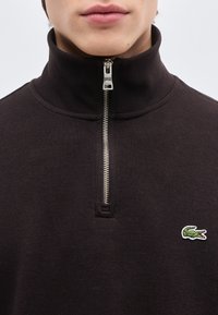 Κοντινό πλάνο ενός ατόμου που φοράει ένα σκουρόχρωμο πουλόβερ με φερμουάρ και ψηλό γιακά, με ένα μικρό πράσινο λογότυπο κροκοδείλου στο στήθος.