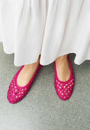 Fødder iført lyserøde flettede flade sko synlige under en hvid knælange nederdel, stående på gråt gulv.