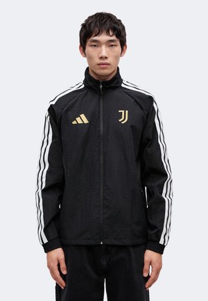 Jeune homme portant une veste de sport noire Adidas Juventus avec des bandes blanches sur les manches, debout devant un fond clair.