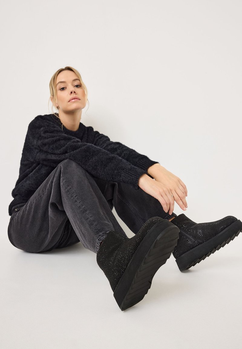 Bottines noires avec des dessus texturés ornés d'accents scintillants, une semelle épaisse et un col rembourré, associées à un pantalon gris ample et un pull.