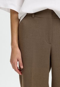 Personne portant un pantalon marron sur mesure et une chemise blanche, la main gauche détendue le long du corps, sur un fond uni.