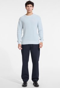Pull en maille côtelée bleu clair avec un col rond, arborant un petit logo rouge. Associé à un pantalon ample bleu marine foncé et des chaussures noires.