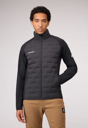 Black puffy jacket with a high collar, quilted design, and long sleeves. Features a logo on the chest and is paired with khaki pants.