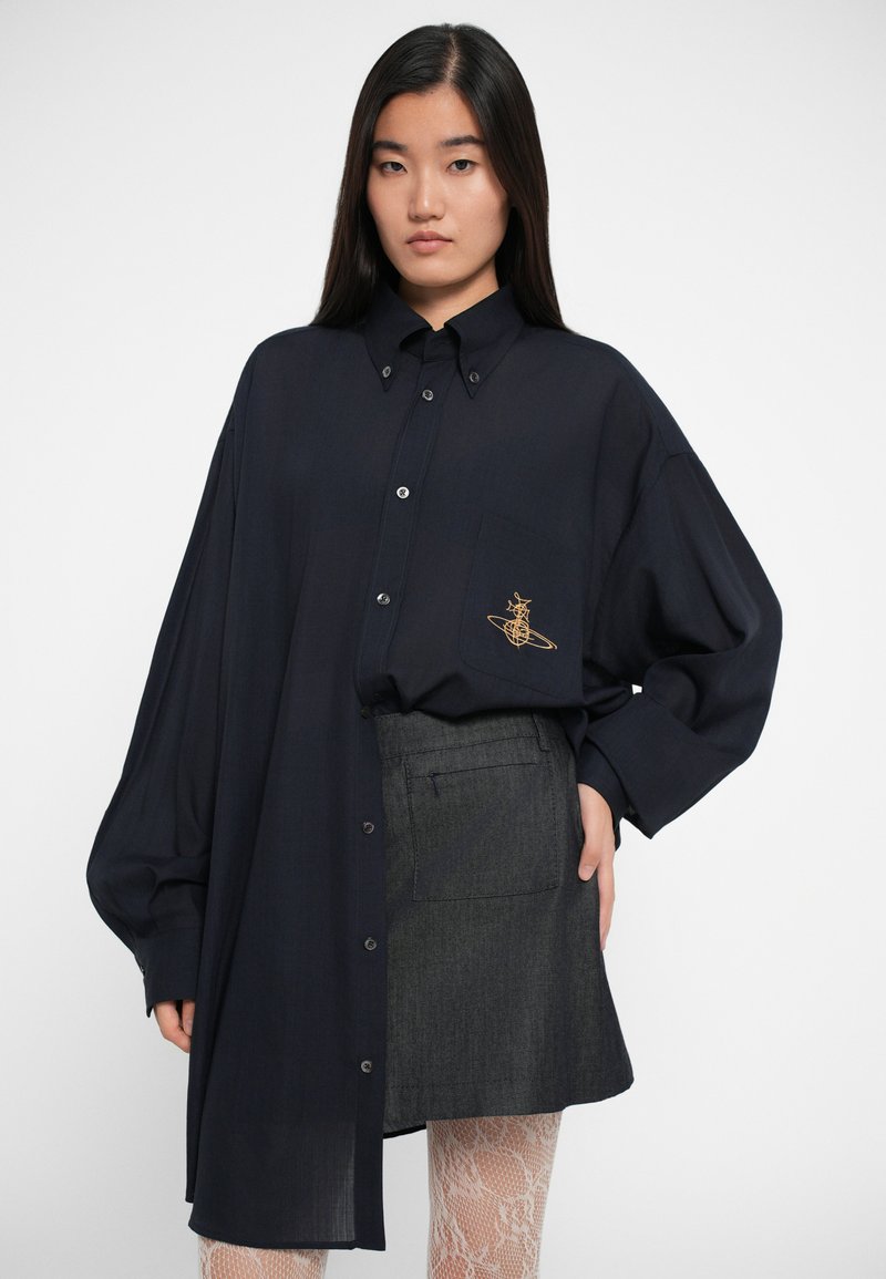 Übergröße navyblaues Hemd mit einer Fronttasche und goldener Stickerei, kombiniert mit einem grauen Rock. Das Hemd hat einen Knopfverschluss und lange Ärmel.