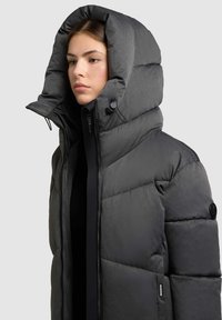 Grå pufferjakke med høj, hættekrave, detaljeret søm og lynlås-lukning. Stoffet har en tekstureret overflade og er isoleret.