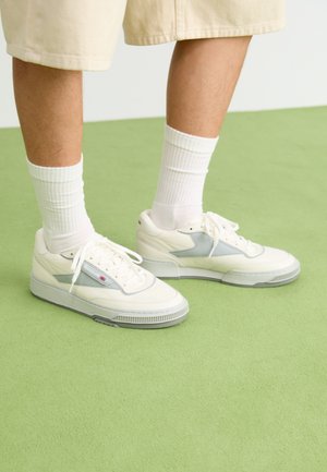 Weiße Ledersneaker mit grauen Akzenten, strukturiertem Zehenbereich und Gummisohle. Getragen mit weißen Knöchelsocken und beigen Shorts auf einer grünen Unterlage.