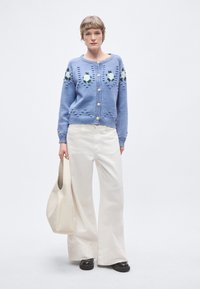 Ανοιχτό μπλε πλεκτό ζακέτα με κεντήματα λουλουδιών, χρυσά κουμπιά και υφασμάτινες λεπτομέρειες, συνδυασμένη με λευκό παντελόνι φαρδιάς γραμμής και μπεζ τσάντα ώμου.