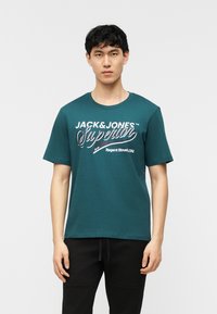Homme portant un t-shirt bleu sarcelle Jack & Jones avec l'inscription "Superior" et un pantalon noir, debout devant un fond uni clair.