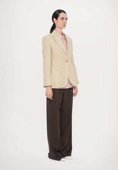 Beige tailored blazer with single button, pink shirt underneath, and wide brown trousers, featuring white pointed-toe shoes.