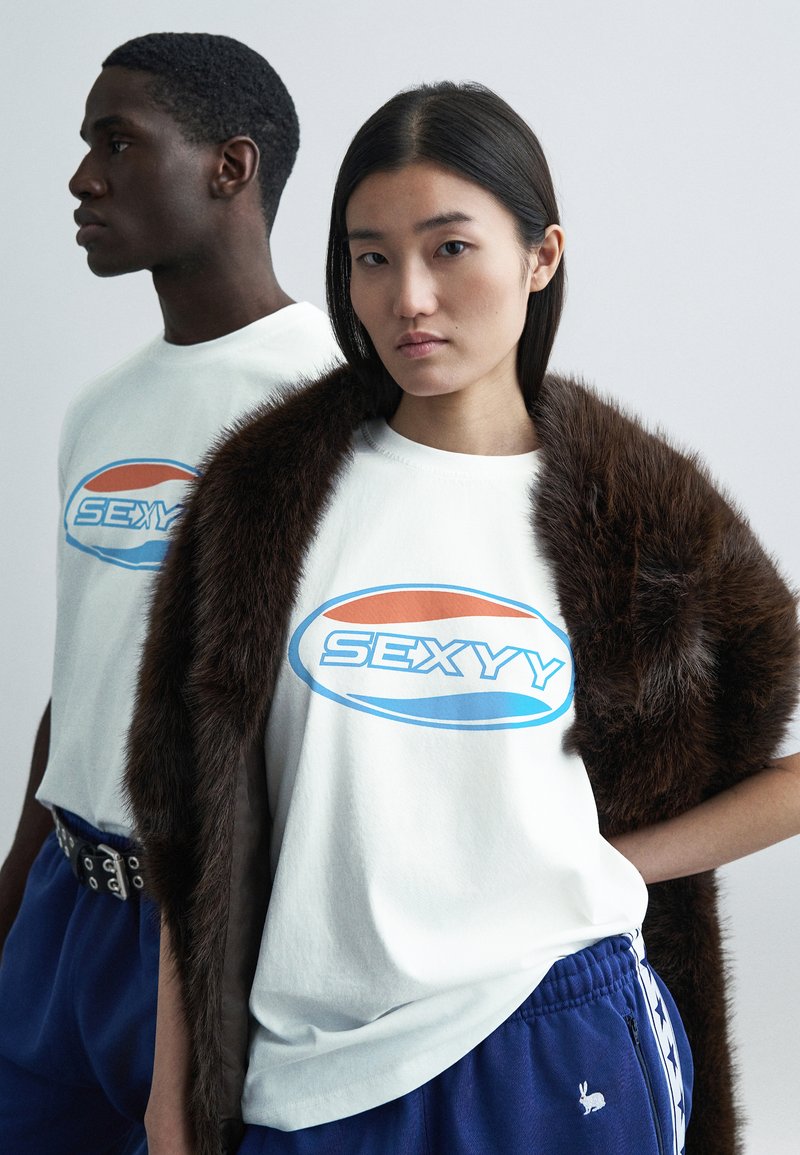 Två modeller bär vita bomullst-shirts med texten "SEXYY" i blått och orange. En modell bär en brun fuskpäls-sjal; båda har blå byxor.