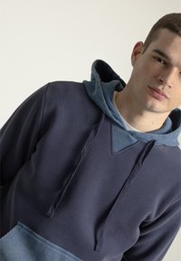 Sudadera con capucha en azul oscuro con una capucha texturizada de un azul más claro. Cuenta con un bolsillo canguro y cordones, fabricada en una tela suave.
