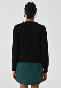 Maglione nero lavorato a maglia con scollatura rotonda e maniche lunghe, con polsini e orlo a coste. Indossato sopra un vestito di raso verde.