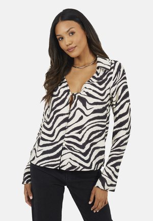 Donna con lunghi capelli scuri che indossa una camicetta con stampa zebrata bianca e nera e pantaloni neri, in piedi con una mano sulla gamba.