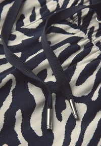 Marinblå och krämfärgad zebra-mönstrad tyger med en strukturerad yta. Svarta dragsnören med silverfärgade metalldetaljer. Nærbild av materialet och designen.