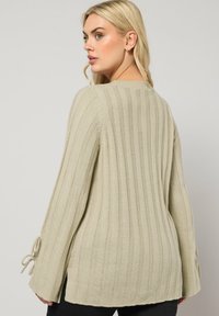 Jasnozielony sweter z prążkowanej tkaniny, z długimi rękawami, bocznymi rozcięciami oraz wiązaniem przy mankietach. Teksturowany splot materiału dodaje głębi.