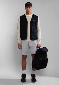 Chaleco navy con múltiples bolsillos sobre un suéter crema, pantalones cortos grises, zapatos negros y una mochila negra, completado con una gorra negra.