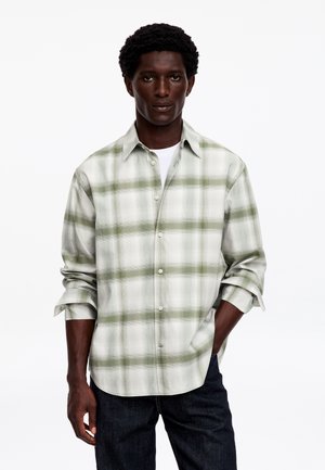 Homme portant une chemise à carreaux vert clair et blanc à boutons, manches retroussées, et un pantalon foncé, debout avec une main dans la poche.