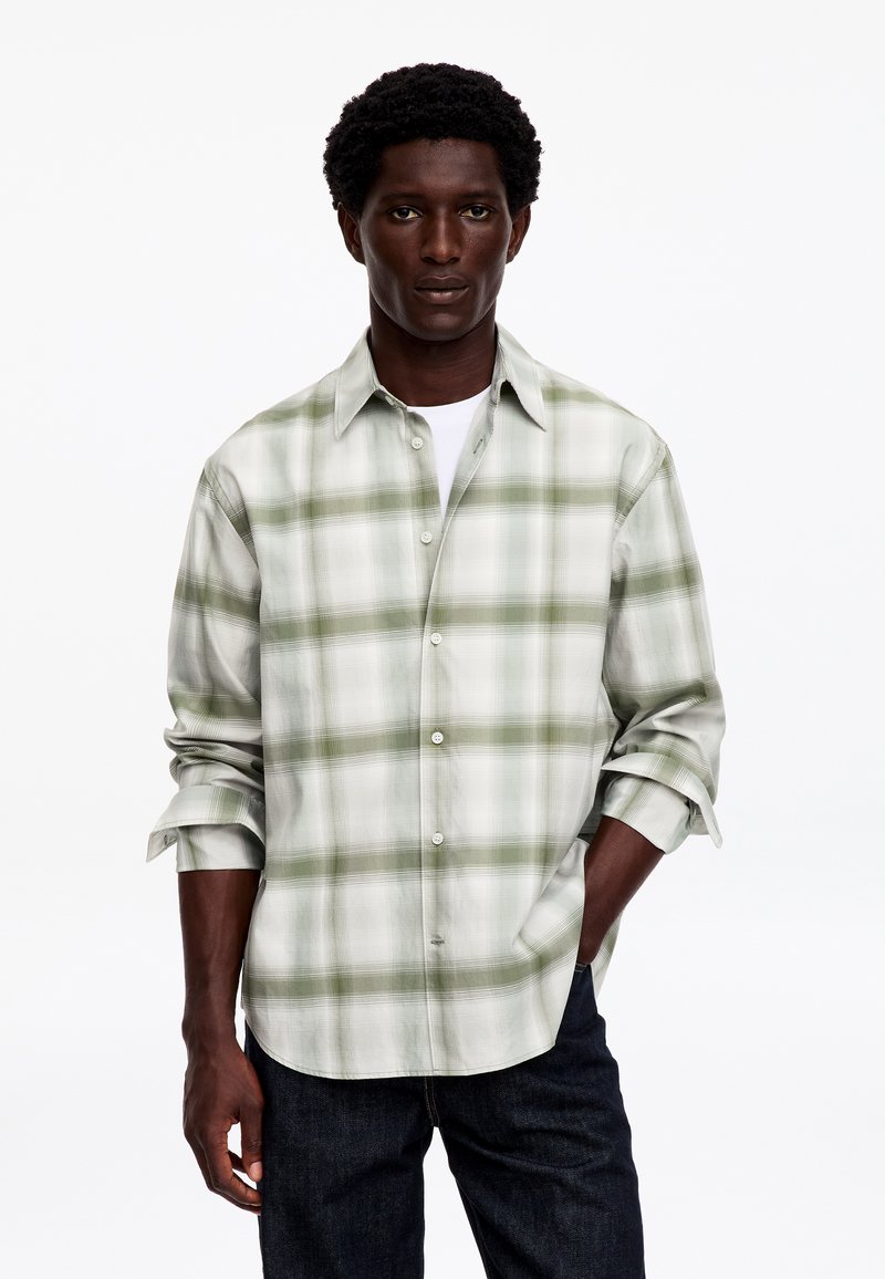Homme portant une chemise à carreaux vert clair et blanc à boutons, manches retroussées, et un pantalon foncé, debout avec une main dans la poche.