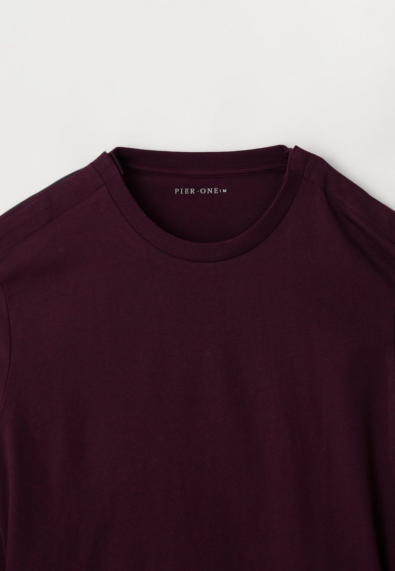 Sötétlila, rövid ujjú crewneck póló lefektetve, mutatja a bels�ő nyak címkét, amelyen a "PIER·ONE" felirat található fehér háttérrel.