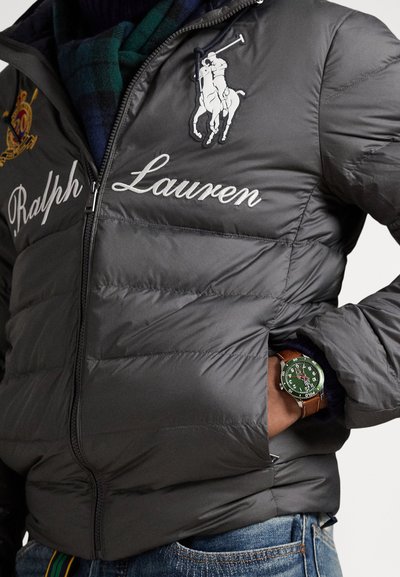Sivá prešívaná bunda s výšivkou "Ralph Lauren", ktorá obsahuje logo koňa, zapínanie na zips a hodinky s zeleným ciferníkom a hnedým remienkom.