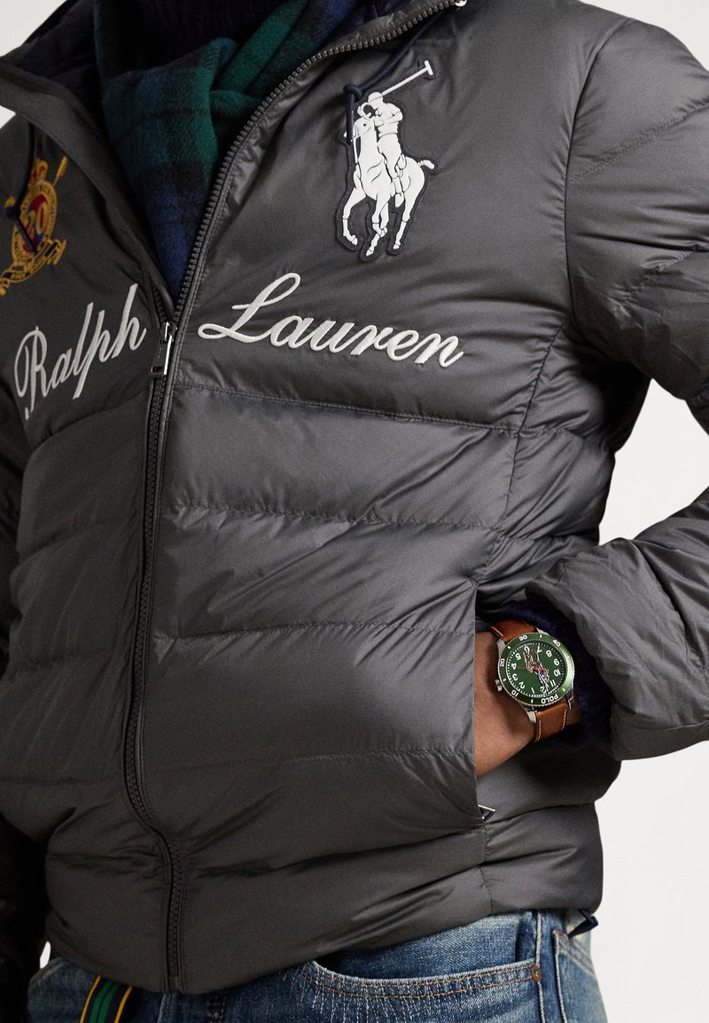Doudoune grise avec broderie "Ralph Lauren" et logo cheval, fermeture éclair, accompagnée d'une montre-bracelet avec cadran vert et bracelet marron.