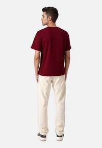 Bordeauxfarbene Baumwoll-T-Shirt mit kurzen Ärmeln, lockerer Passform, kombiniert mit cremefarbener Hose und dunklen Sneakers, von hinten gesehen.