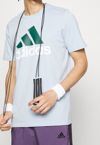 Světle modré tričko Adidas s bílým logem a zelenými akcenty, doplněné fialovými šortkami. Černá skákací šňůra s texturovanými úchyty držena v rukou.