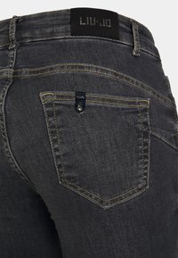 Jeans in denim grigio scuro con un design aderente. Caratteristiche includono cuciture gialle, un'etichetta nera brandizzata e una piccola tasca sul retro con accessori.