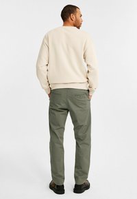 Beige Crewneck-Sweatshirt mit gerippten Bündchen, kombiniert mit olivgrünen Chinos mit zwei Gesäßtaschen und getragen mit schwarzen Schuhen.