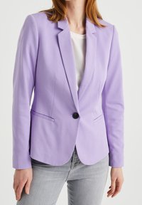 Žena ve fitted levandulovém blejzru přes bílé tričko a světle šedých džínách, stojící na pozadí s čistě bílou barvou.