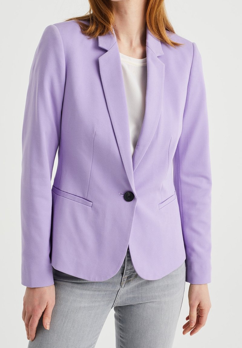 Žena ve fitted levandulovém blejzru přes bílé tričko a světle šedých džínách, stojící na pozadí s čistě bílou barvou.