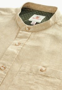 Chemise beige à col avec un motif texturé, comportant deux boutons et une poche avant. Le col intérieur présente un motif de vagues vertes.