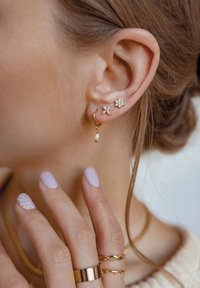 Orecchini a cerchio in oro con goccia di perla, orecchini a perno floreali con gemme trasparenti, e una mano adornata con più anelli d'oro e unghie di un leggero colore viola.