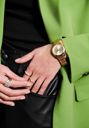 Guldring med en rund urskive, metallisk rem og en beige skive. Båret på en hånd med flere ringe og en grøn blazer over en sort top.